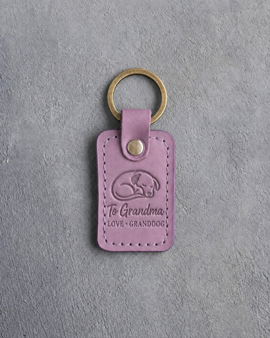 Lilac leather keychain debossed with a curled sleeping dog above To Grandma and LOVE · GRANDDOG, on gray concrete