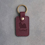 Sangria red leather keychain debossed with a Shih Tzu silhouette and the words Topknot ROYALTY, on gray concrete