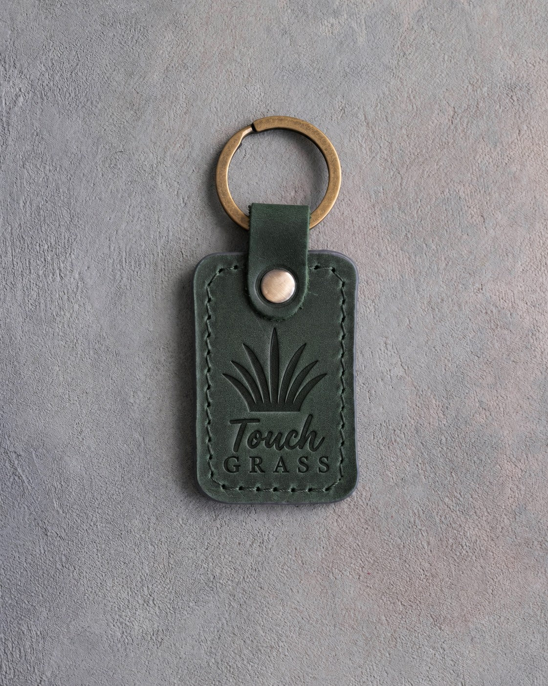 Forest green leather keychain debossed with a tuft of grass above Touch GRASS, on gray concrete