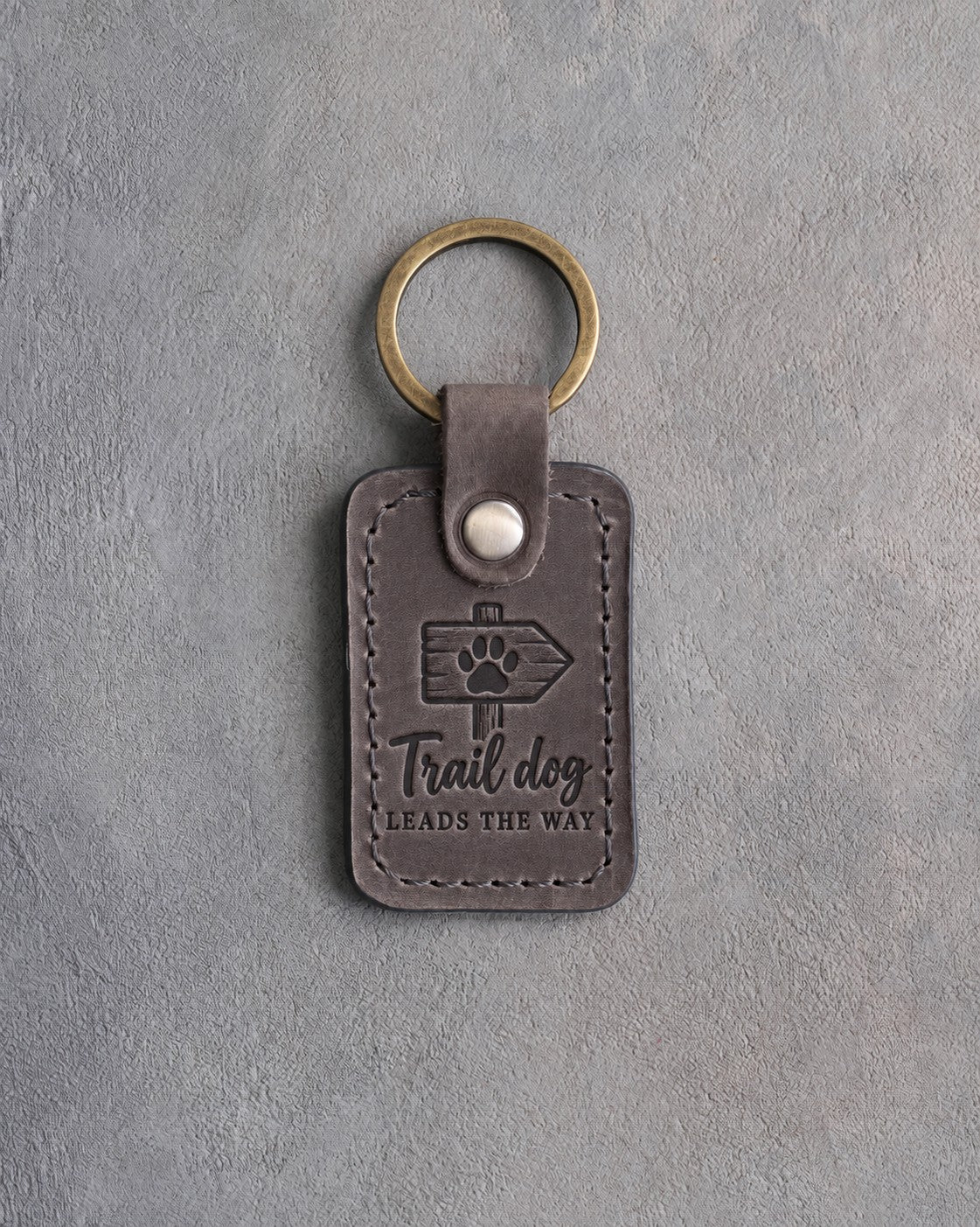 Country gray leather keychain debossed with a trail marker arrow and a paw above the words Trail dog LEADS THE WAY, on gray concrete