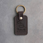 Dark espresso leather keychain debossed with a coiled leash and the words Trains the HUMAN END, on gray concrete
