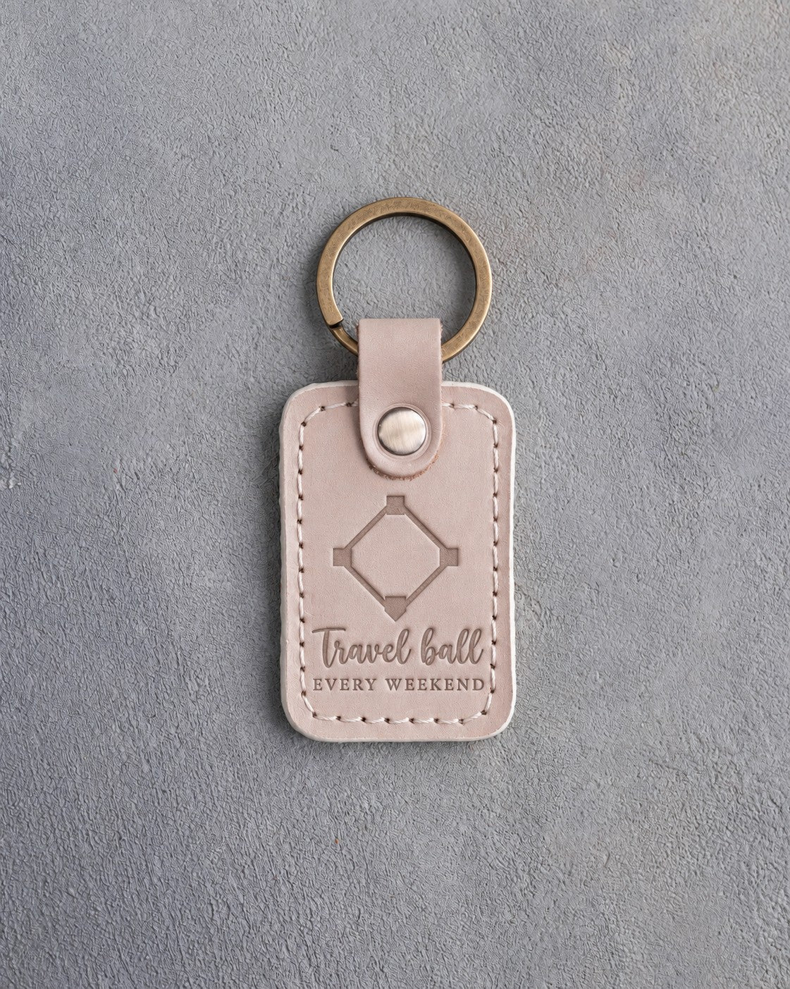 Gray sand leather keychain debossed with a baseball diamond and the words Travel ball EVERY WEEKEND, on gray plaster