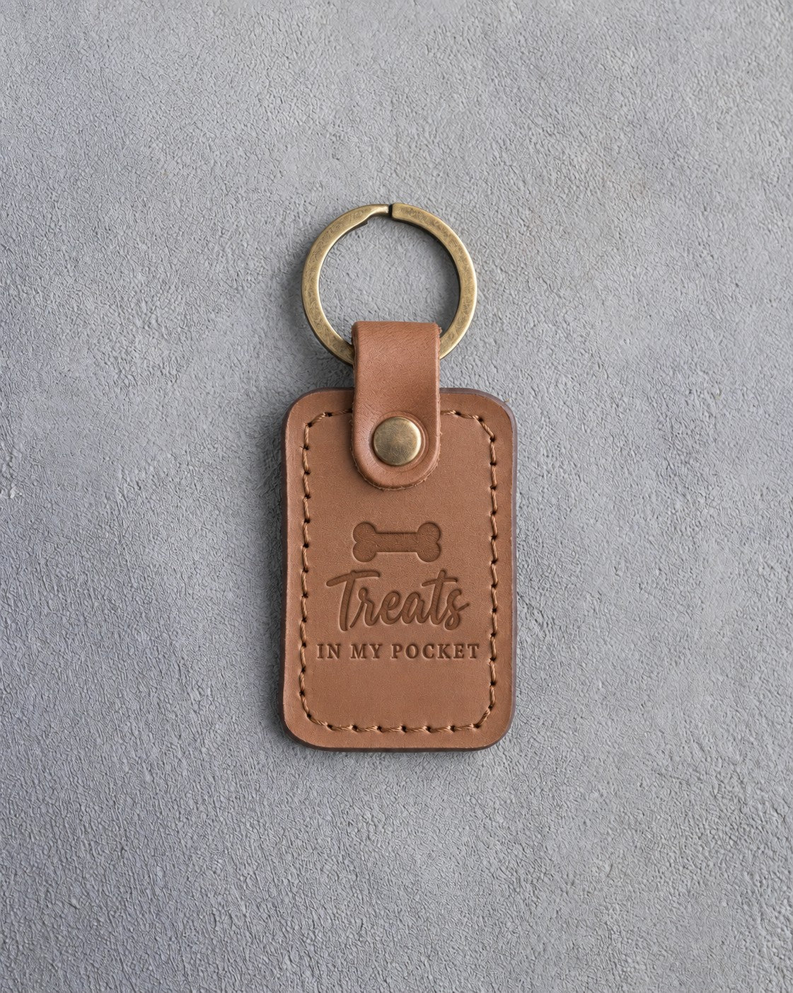 Caramel leather keychain debossed with a bone-shaped dog biscuit and the words Treats IN MY POCKET, on gray concrete