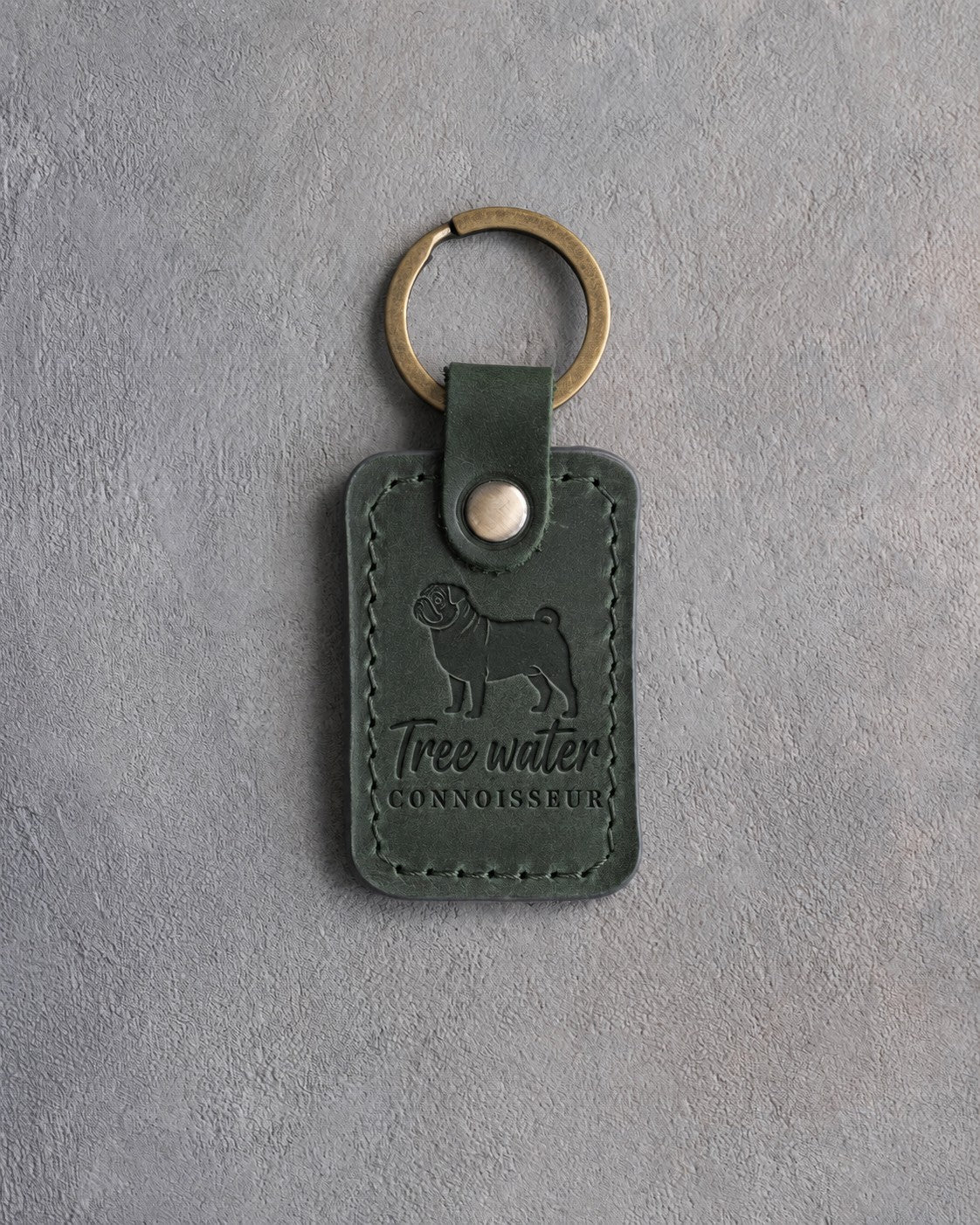 Forest green leather keychain debossed with a pug silhouette and the words Tree water CONNOISSEUR, on gray concrete