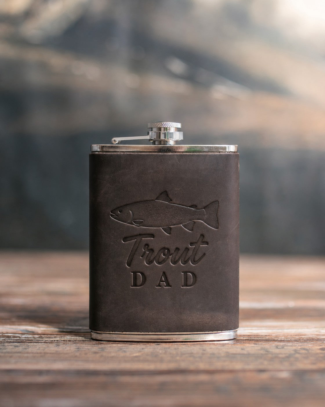 Dark espresso leather-wrapped hip flask debossed with a rainbow trout above the words Trout DAD, on a wooden table
