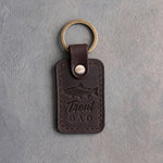 Dark espresso leather keychain debossed with a rainbow trout above the words Trout DAD, on gray plaster