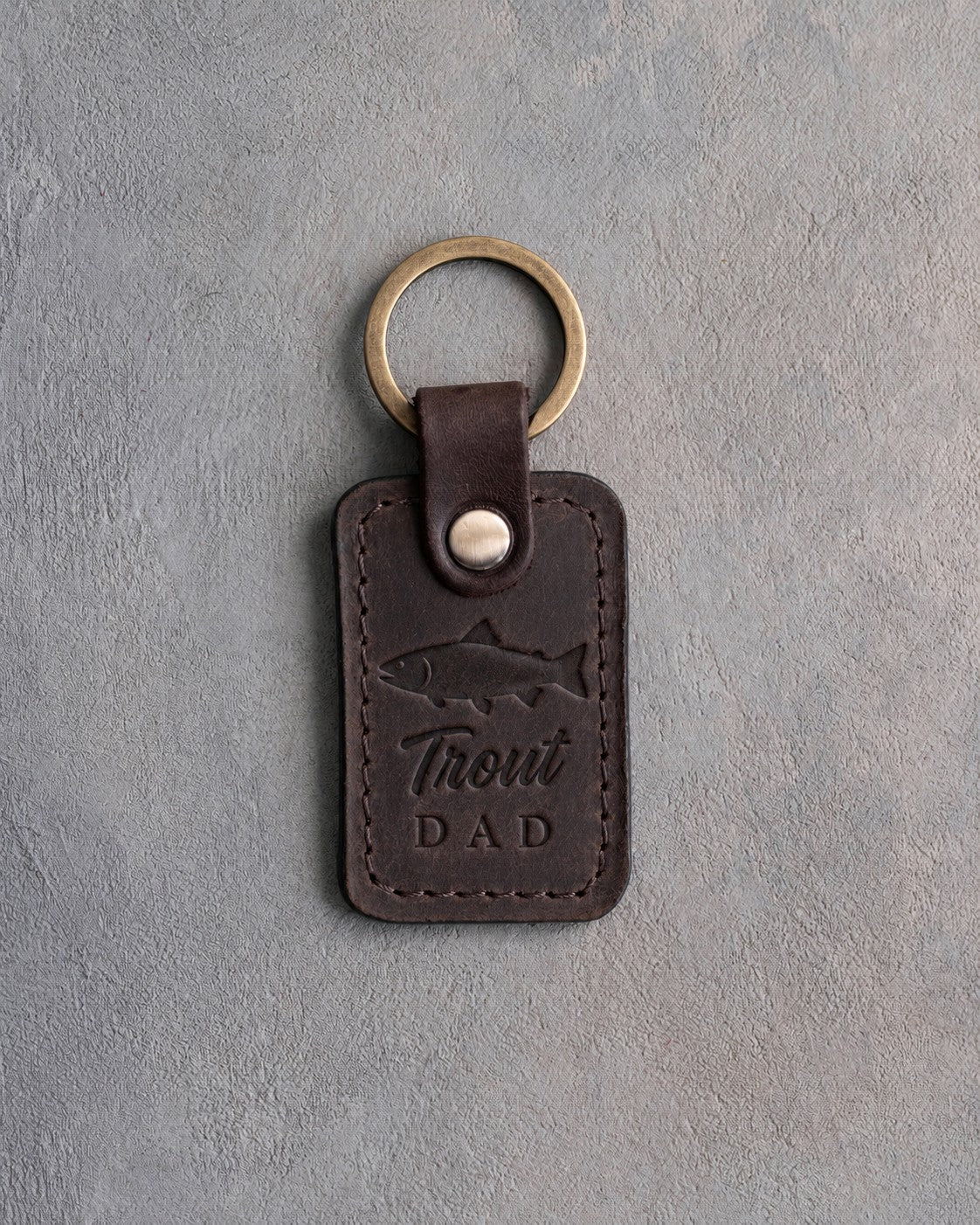Dark espresso leather keychain debossed with a rainbow trout above the words Trout DAD, on gray plaster