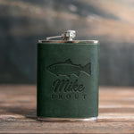 Forest green leather-wrapped hip flask debossed with a rainbow trout above Mike and TROUT, on a wooden table