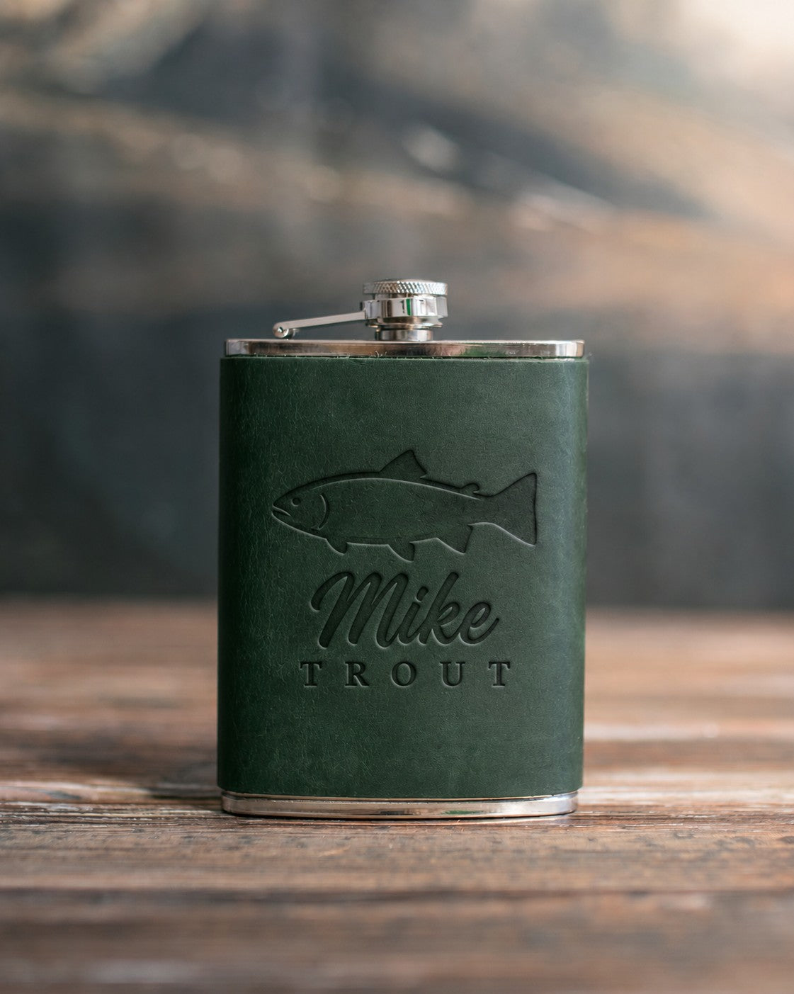 Forest green leather-wrapped hip flask debossed with a rainbow trout above Mike and TROUT, on a wooden table