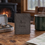 Country gray Owen FLY FISHING card wallet in full-grain leather on a knotty pine cabin table beside an open fly box and a green enamel coffee mug