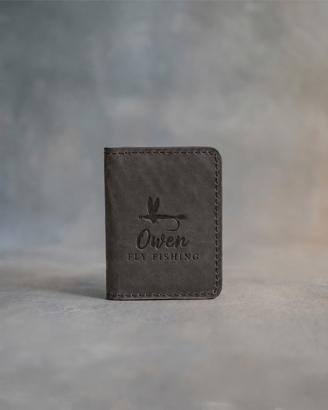 Country gray leather card wallet standing on gray plaster, debossed with a dry fly above Owen and FLY FISHING