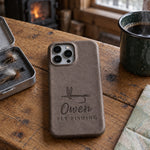 Country gray Owen FLY FISHING iPhone case in full-grain leather on a knotty pine cabin table beside an open fly box and a green enamel coffee mug