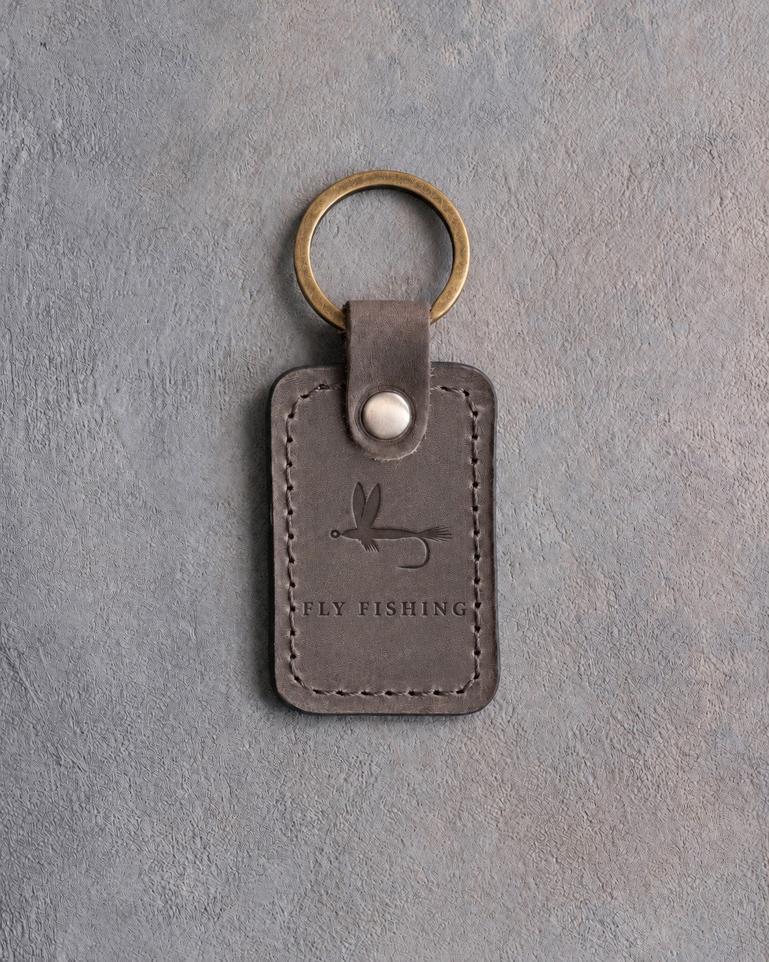 Country gray leather keychain debossed with a dry fly above FLY FISHING, on gray concrete