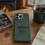 Forest green Mike TROUT iPhone case in full-grain leather on a knotty pine cabin table beside an open fly box and a green enamel coffee mug