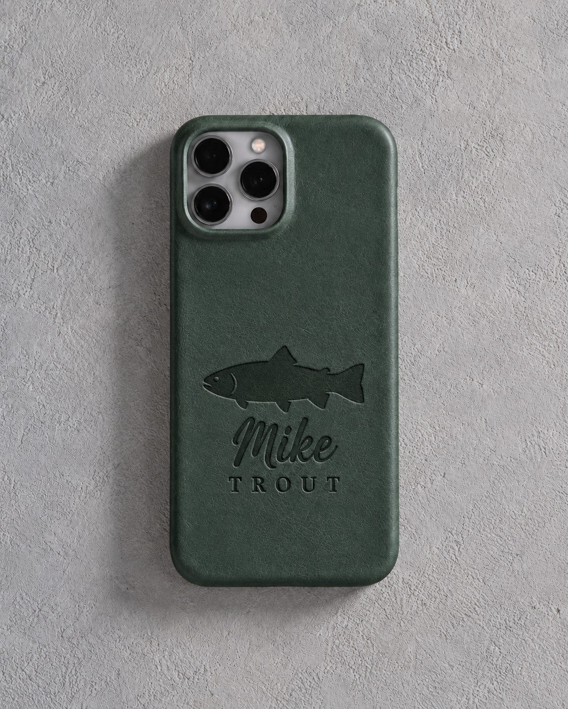 Back of a forest green leather iPhone case debossed with a rainbow trout above Mike and TROUT below the camera, on gray plaster