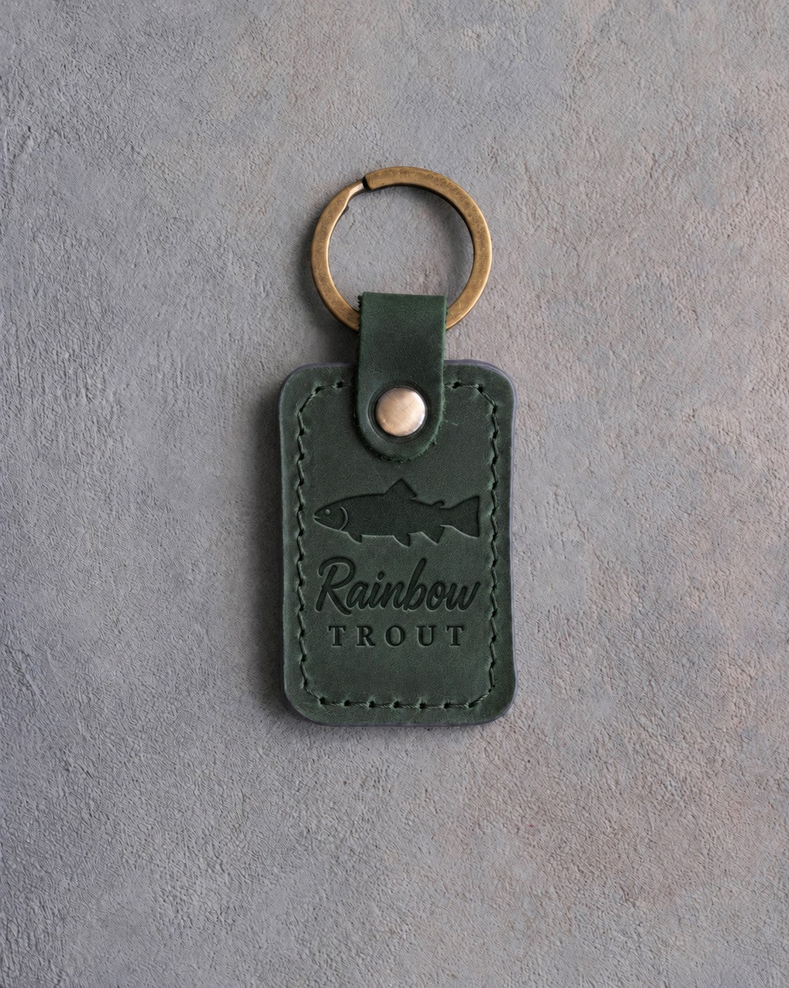 Forest green leather keychain debossed with a rainbow trout above Rainbow and TROUT, on gray concrete