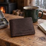 Dark espresso Mike TROUT bifold wallet in full-grain leather on a knotty pine cabin table beside an open fly box and a green enamel coffee mug