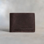 Closed dark espresso leather bifold wallet with a rainbow trout beside Mike and TROUT pressed in the lower right corner, on gray plaster