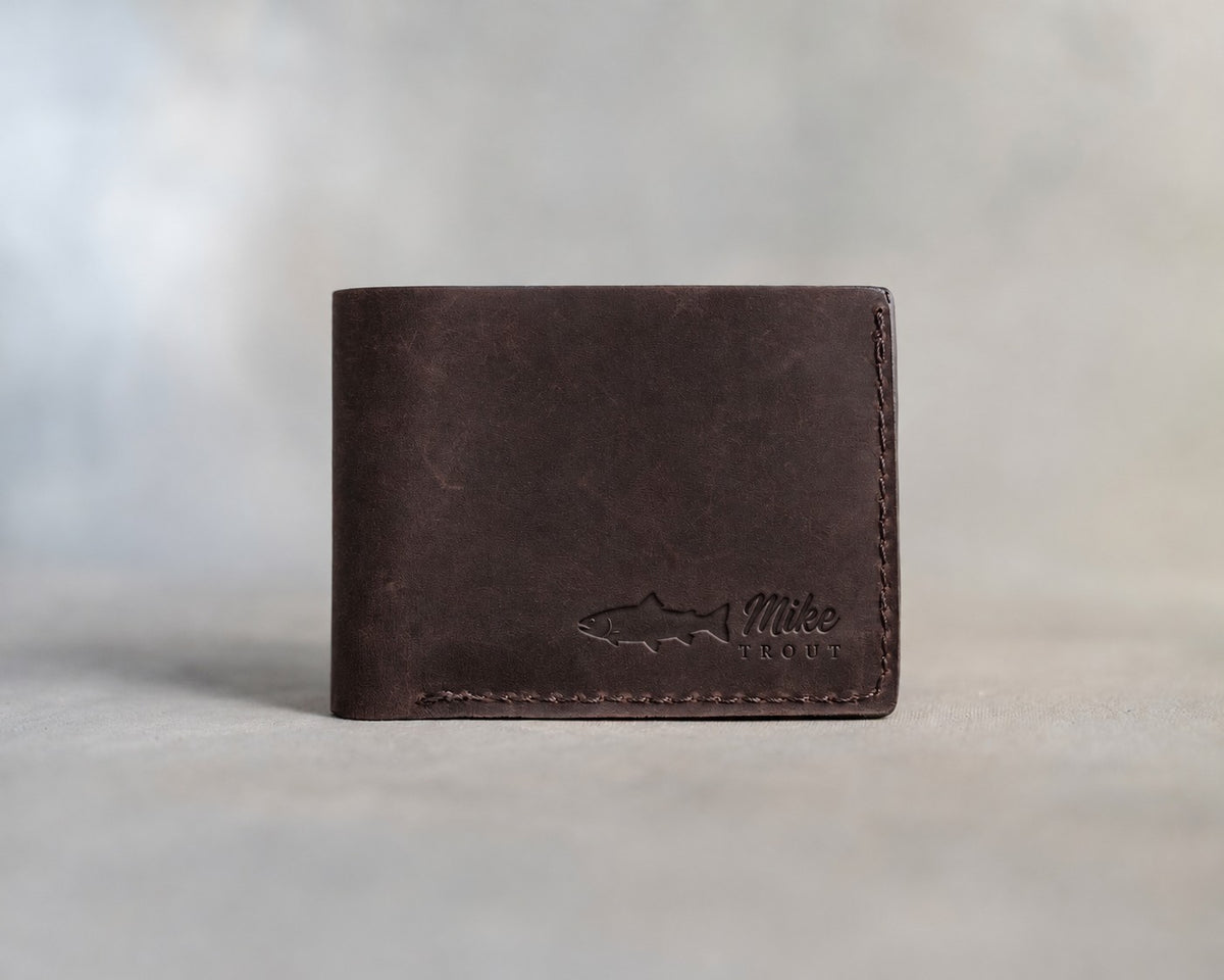 Closed dark espresso leather bifold wallet with a rainbow trout beside Mike and TROUT pressed in the lower right corner, on gray plaster