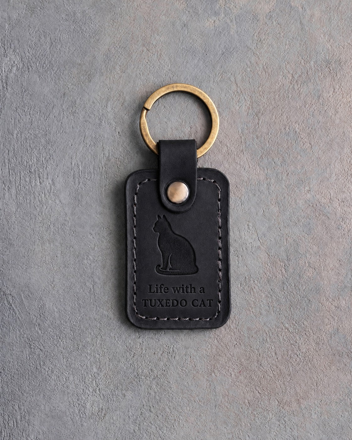 Black leather keychain with a debossed sitting cat above Life with a and TUXEDO CAT, on gray concrete