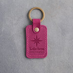 Fuchsia leather keychain debossed with an eight-point compass star above the words Twelve homes ONE FAMILY, on gray concrete