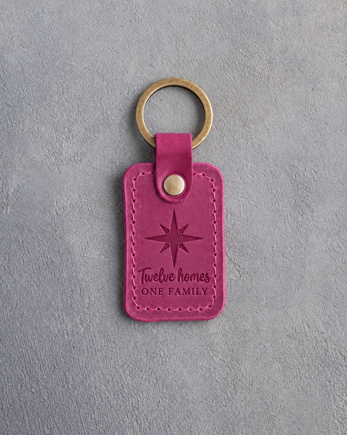Fuchsia leather keychain debossed with an eight-point compass star above the words Twelve homes ONE FAMILY, on gray concrete