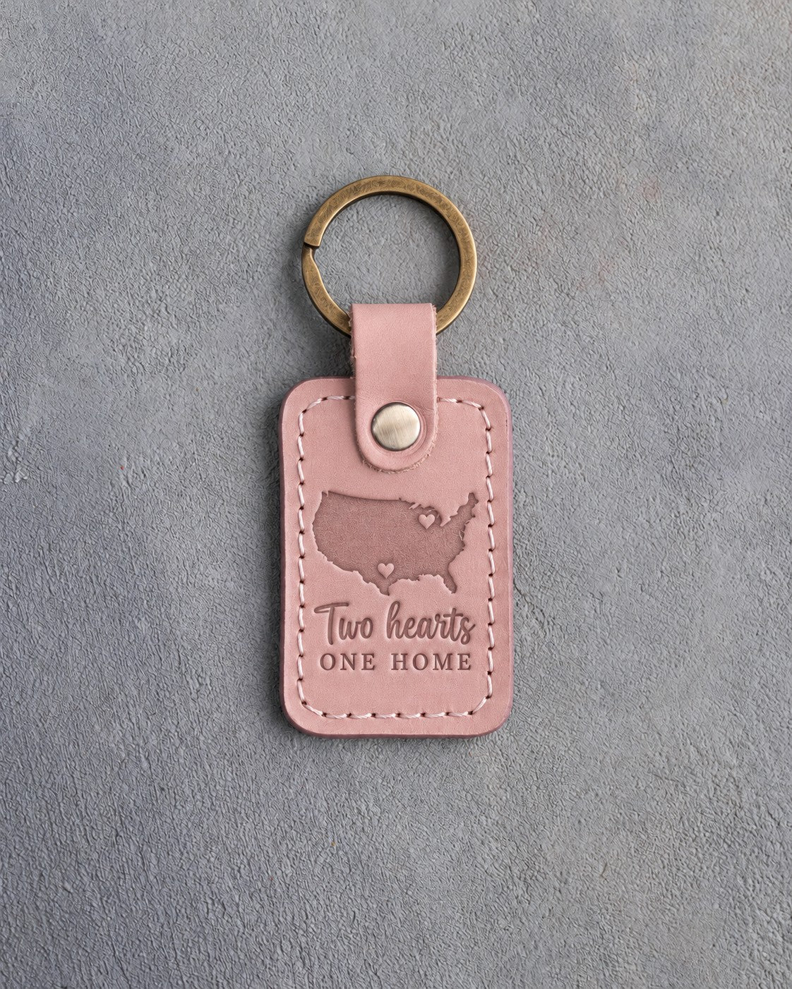 Blush pink leather keychain debossed with a map of the lower forty-eight states and two hearts above Two hearts and ONE HOME, on gray concrete