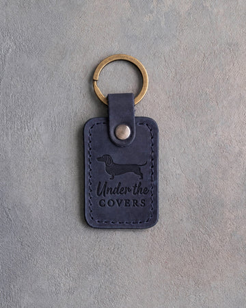 Blueberry navy leather keychain debossed with a dachshund silhouette and the words Under the COVERS, on gray concrete