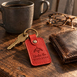 Under the MISTLETOE keychain in bright red leather on an oak kitchen table beside a worn wallet and two stoneware mugs