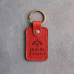 Poppy red leather keychain debossed with a sprig of mistletoe above the words Under the MISTLETOE, on gray concrete