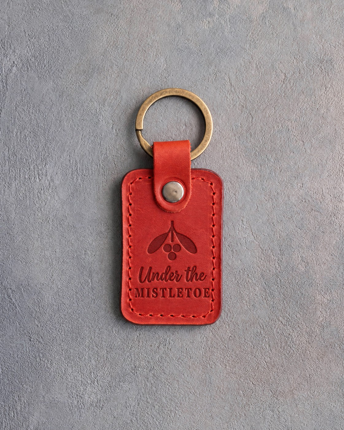 Poppy red leather keychain debossed with a sprig of mistletoe above the words Under the MISTLETOE, on gray concrete
