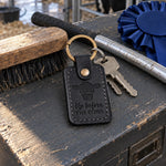 Black Up Before the Cows keychain on a painted show box lid in a livestock barn alley beside a show stick and a blue rosette