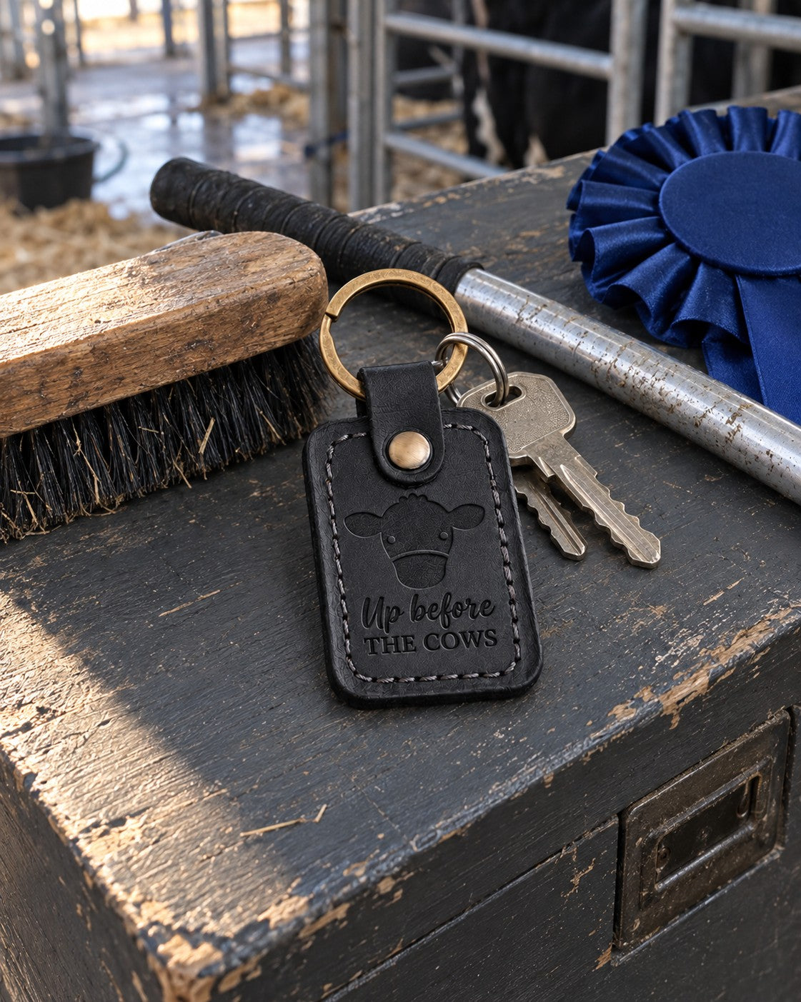 Black Up Before the Cows keychain on a painted show box lid in a livestock barn alley beside a show stick and a blue rosette