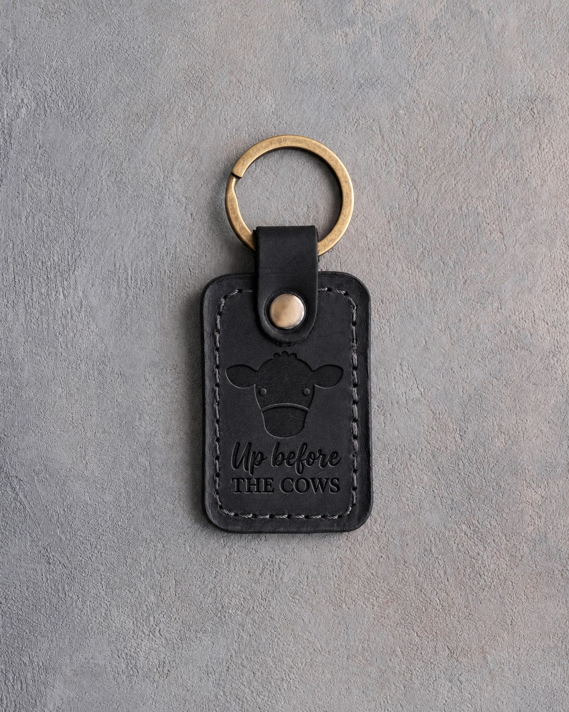 Black leather keychain debossed with a front-facing cow's face above the words Up before THE COWS, on gray concrete