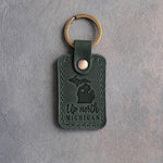 Forest green leather keychain debossed with Michigan's two peninsulas and a heart at Traverse City above Up north and MICHIGAN, on gray concrete