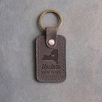 Country gray leather keychain debossed with a New York State silhouette and a heart at Syracuse above Upstate and NEW YORK, on gray concrete