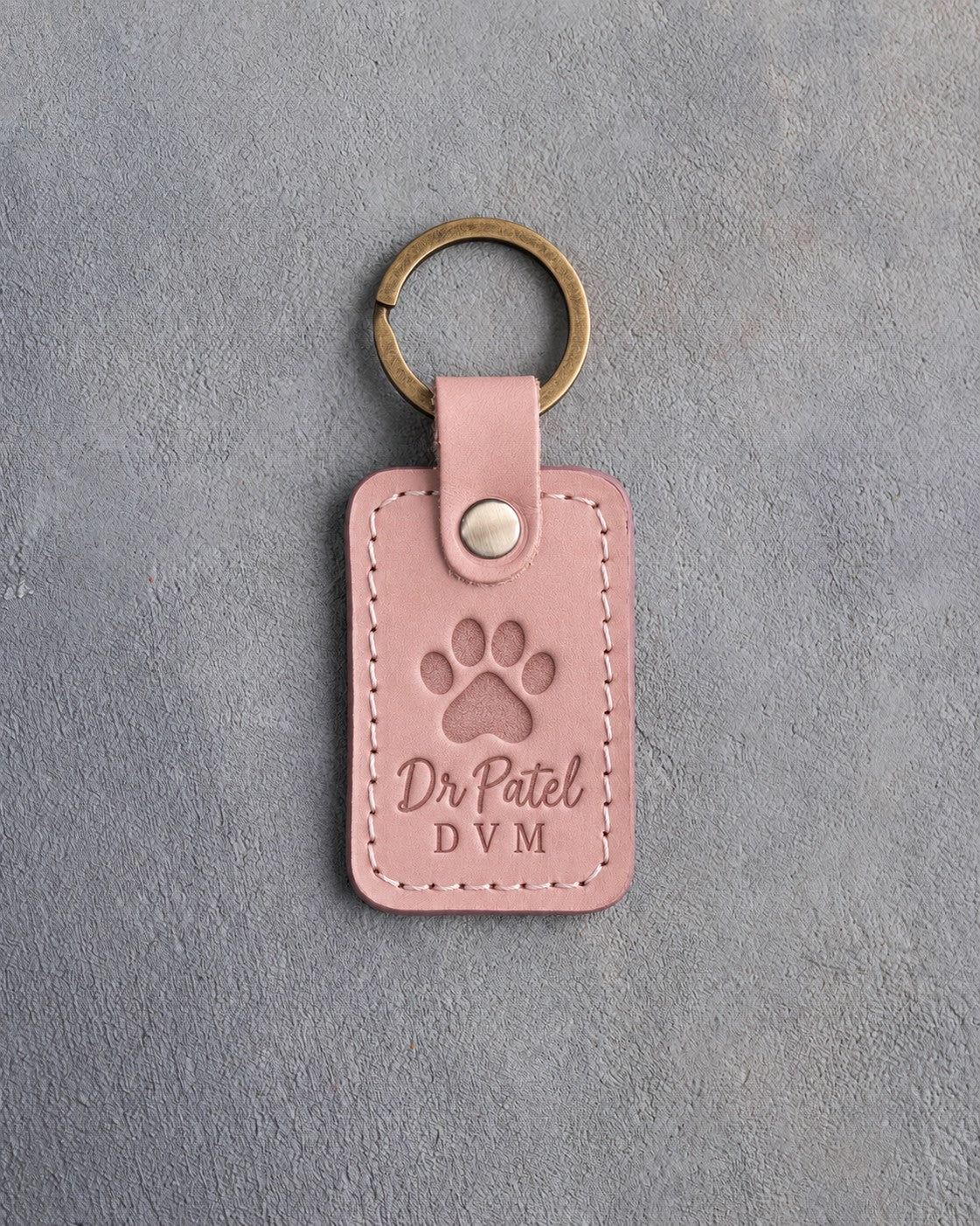 Blush pink leather keychain debossed with a paw print above the name Dr Patel and DVM, on gray plaster