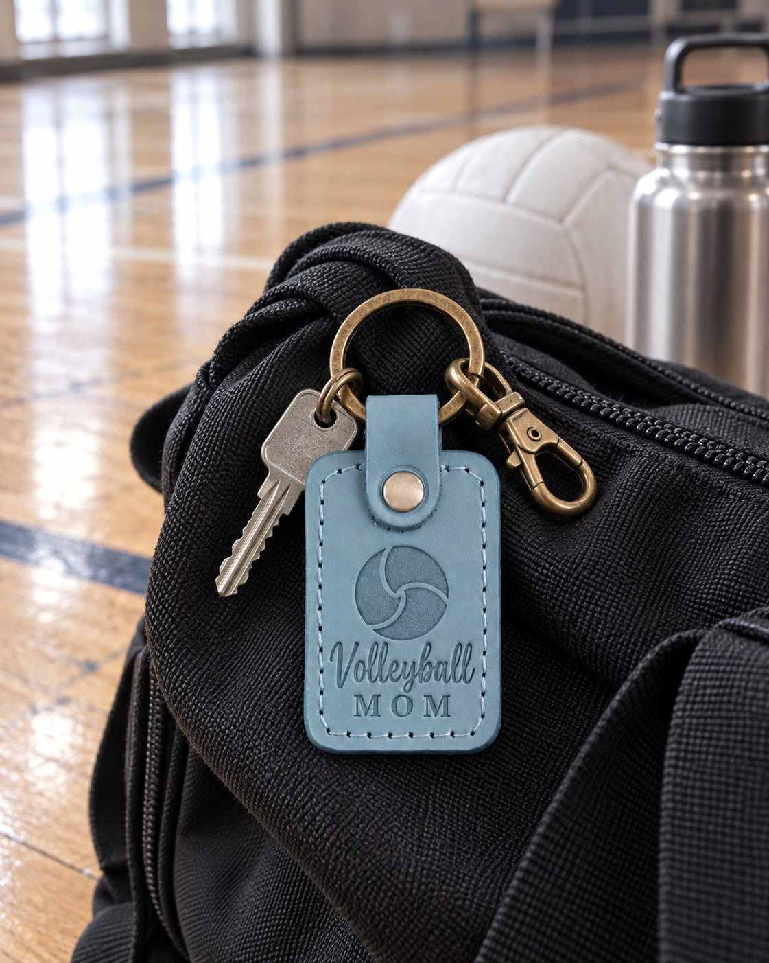 Turquoise Volleyball Mom keychain clipped to a backpack beside a volleyball in a tournament gym