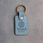 Turquoise leather keychain debossed with a volleyball and the words Volleyball MOM, on gray plaster