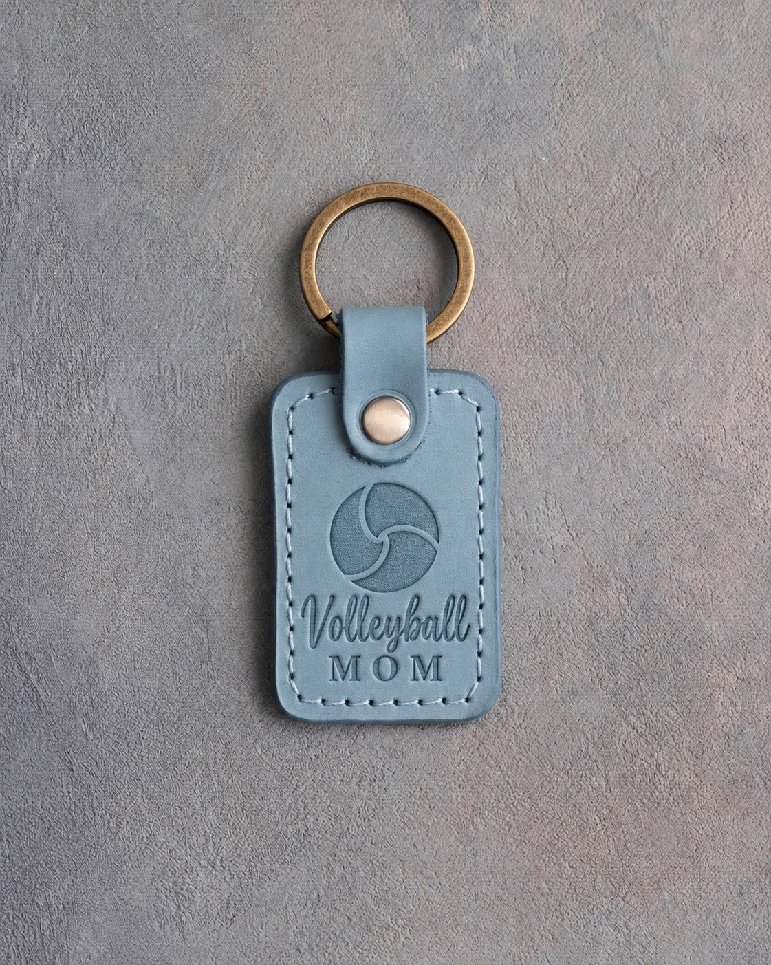 Turquoise leather keychain debossed with a volleyball and the words Volleyball MOM, on gray plaster