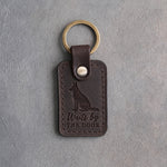 Dark espresso leather keychain debossed with a German shepherd sitting at attention and the words Waits by THE DOOR, on gray concrete