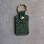 Forest green leather keychain debossed with a looping dog leash above The dog and WALKS DAD, on gray concrete