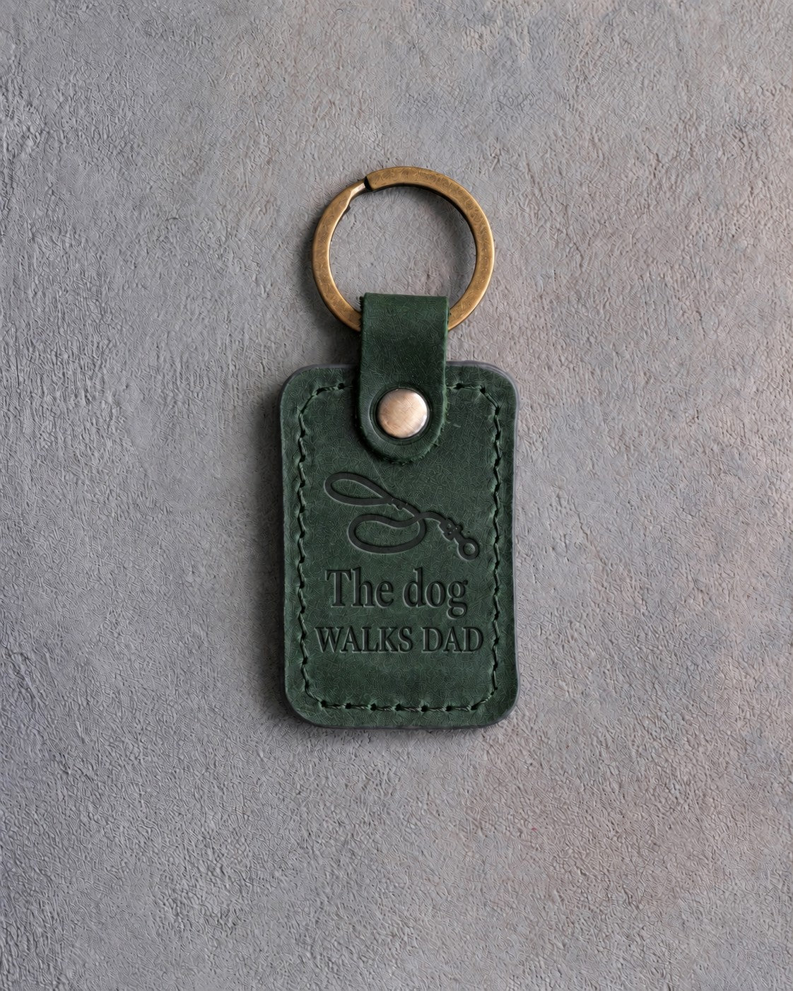 Forest green leather keychain debossed with a looping dog leash above The dog and WALKS DAD, on gray concrete
