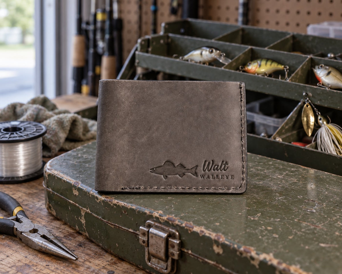 Country gray Walt WALLEYE bifold wallet in full-grain leather on the lid of an olive-green steel tackle box in a garage, lures in the trays behind