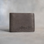 Closed country gray leather bifold wallet with a walleye beside Walt and WALLEYE pressed in the lower right corner, on gray plaster