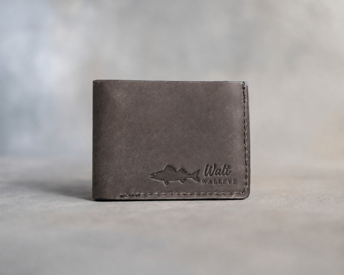 Closed country gray leather bifold wallet with a walleye beside Walt and WALLEYE pressed in the lower right corner, on gray plaster