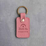 Parisian blossom pink leather keychain debossed with a fermata above the words Watch the DIRECTOR, on gray plaster