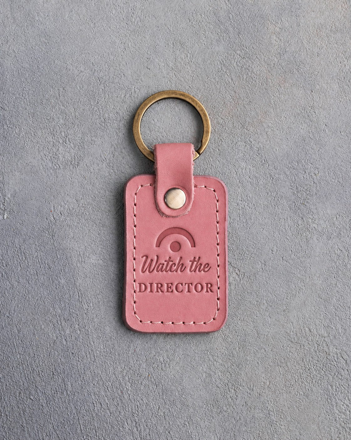 Parisian blossom pink leather keychain debossed with a fermata above the words Watch the DIRECTOR, on gray plaster
