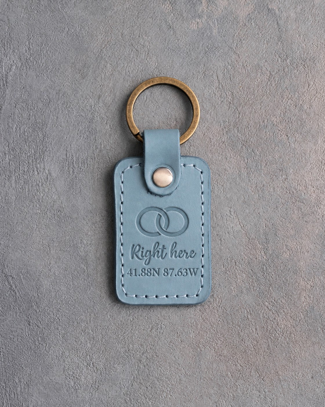 Turquoise leather keychain debossed with two interlocked wedding rings above Right here and 41.88N 87.63W, on gray concrete