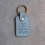 Turquoise leather keychain debossed with two interlocked wedding rings above Right here and WITH YOU, on gray concrete